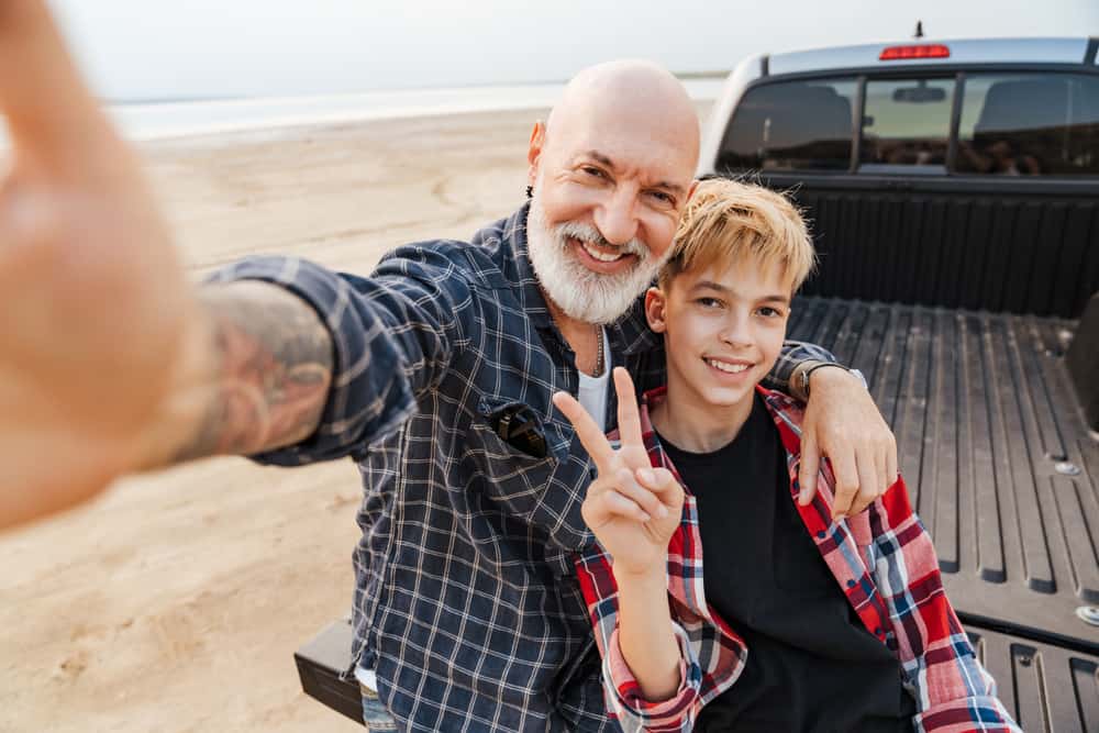 Smiling mid aged father and his preschooler son sitting on a back of a car parked at the beach taking a selfie